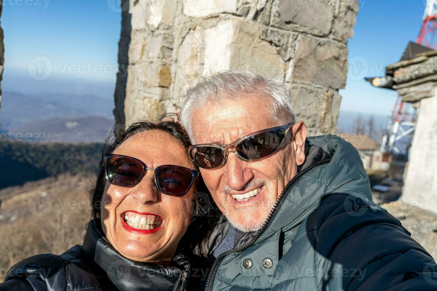 middle aged couple wearing winter clothes taking a selfie over a mountain photo