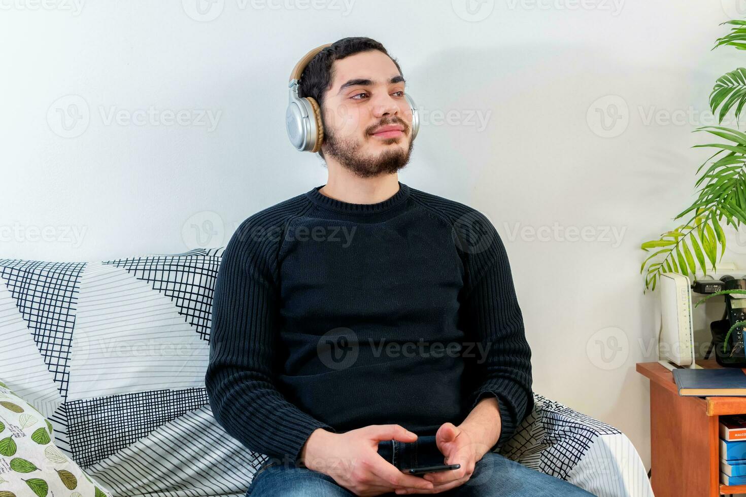 Front view of a happy man with headphones using a mobile phone on line sitting on a couch photo