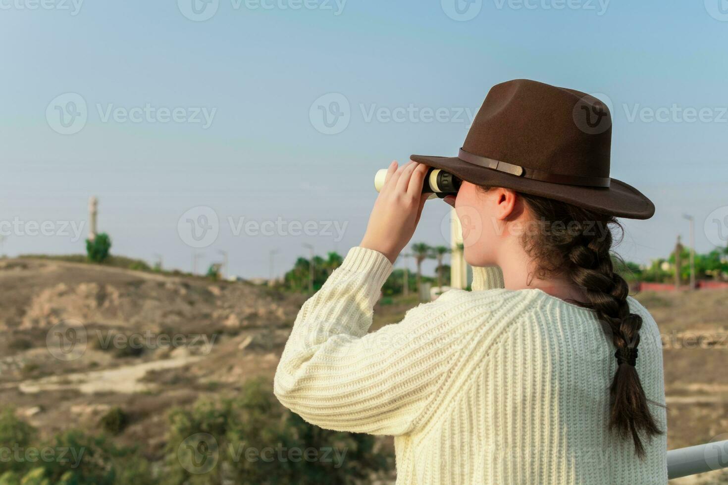 Young woman with brown hat and beige sweater, looking through binoculars, in natural park. photo