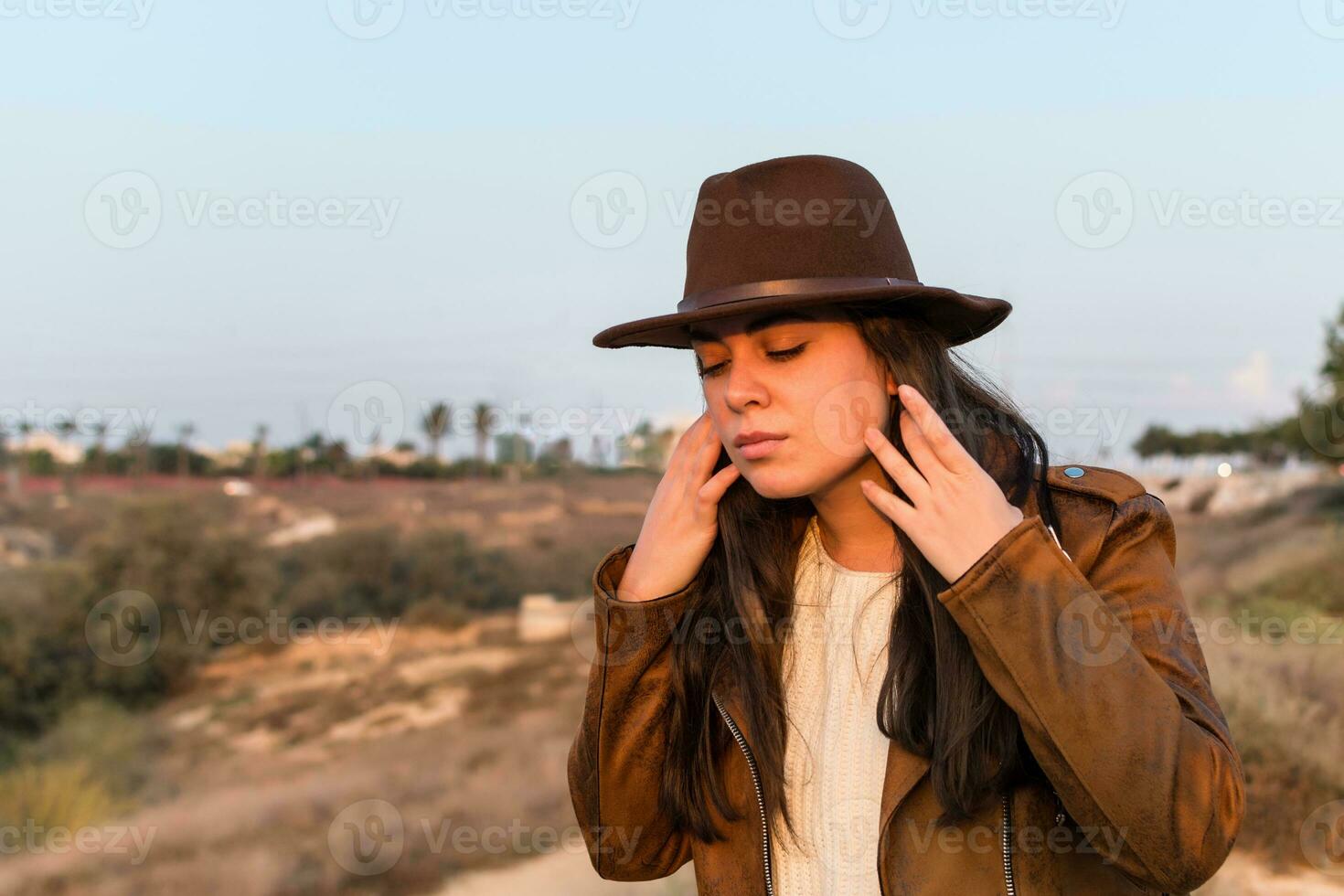 Young woman with hat and brown leather jacket, straightens her hair, in natural park. photo