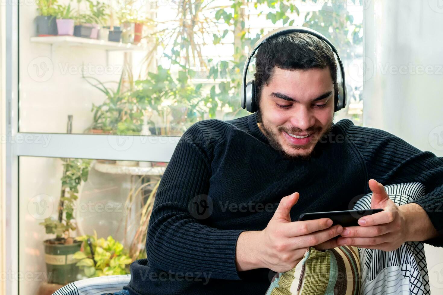 Front view of a happy man with headphones using a mobile phone on line sitting on a couch photo