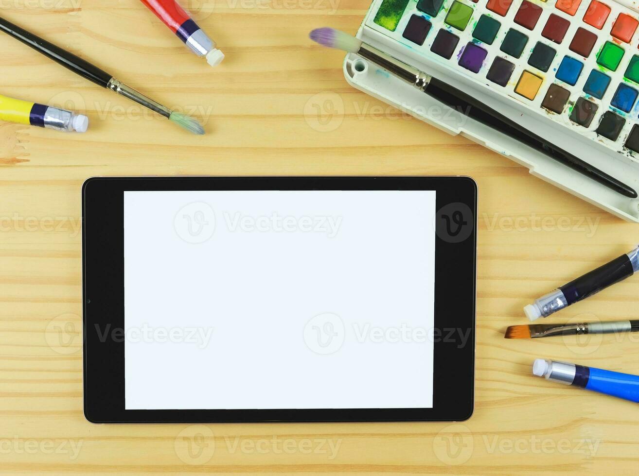 flat lay of artist workspace with digital tablet. Drawing tools, stationary supplies, Watercolor paints, brushes and white blank screen of digital tablet on wooden table, copy space. photo