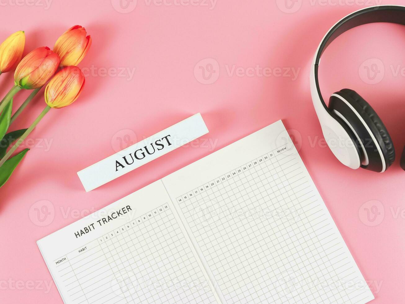 flat lay of habit tracker book with wooden calendar August,  headphones and tulips  on pink background. photo