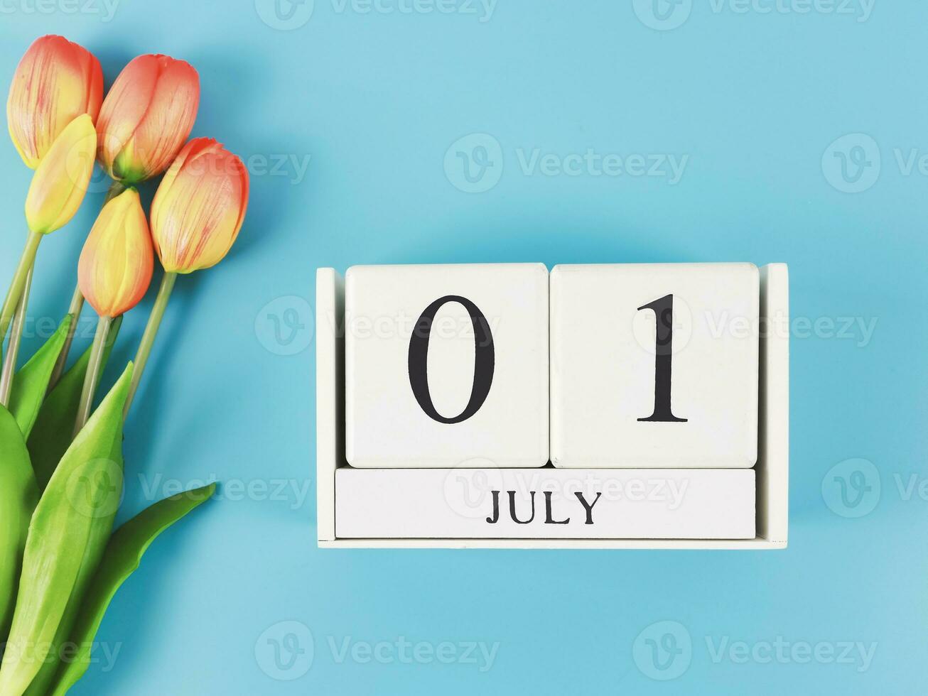 flat lay of wooden calendar with date July 01 on blue  background with orange and yellow tulips, copy space. photo