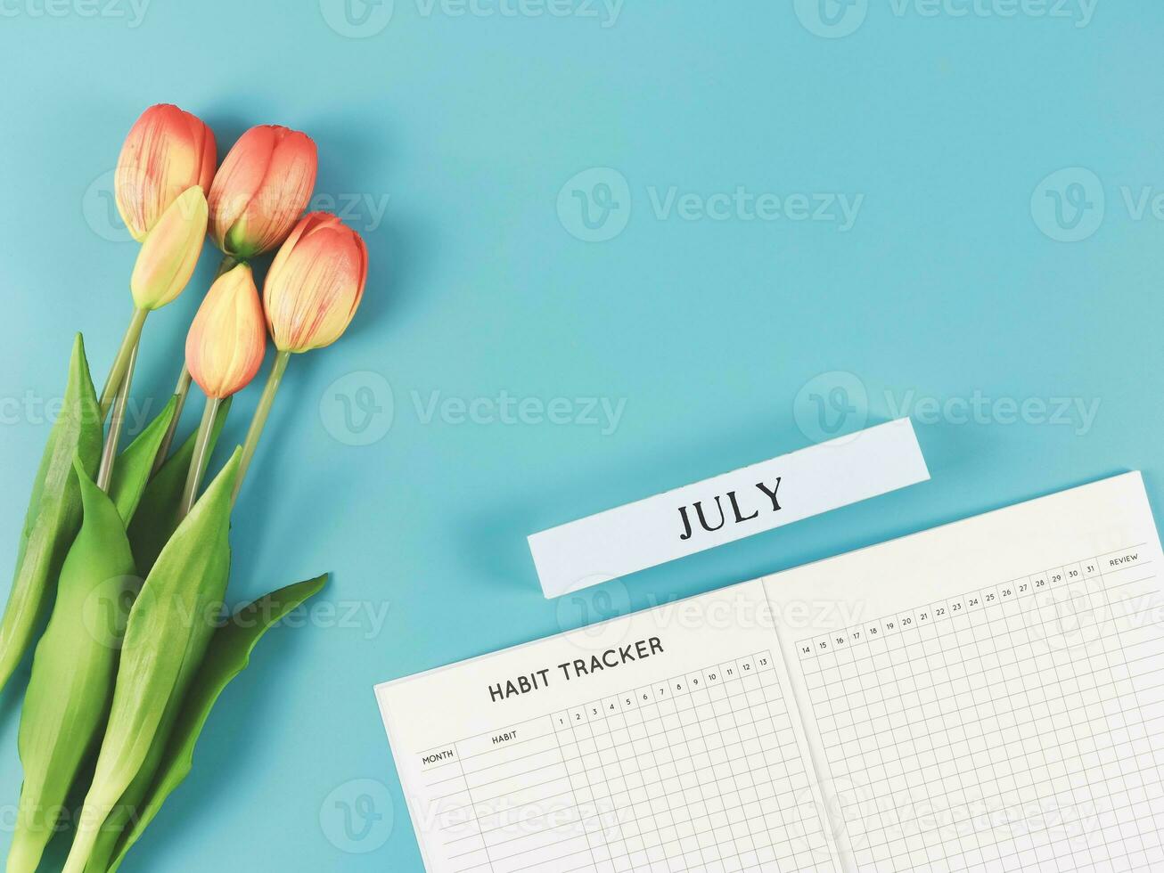 flat lay of habit tracker book with wooden calendar July,   and tulips  on blue background. photo