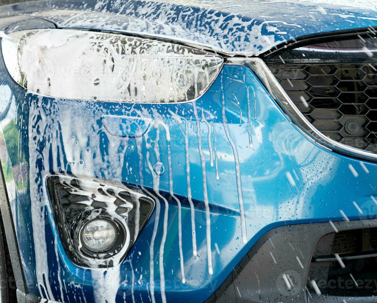 Blue car wash with white soap foam. Auto care business. Car cleaning service. Vehicle cleaning service. Foam wash car detailing. Headlamp light of the blue luxury SUV car is covered with white foam. photo