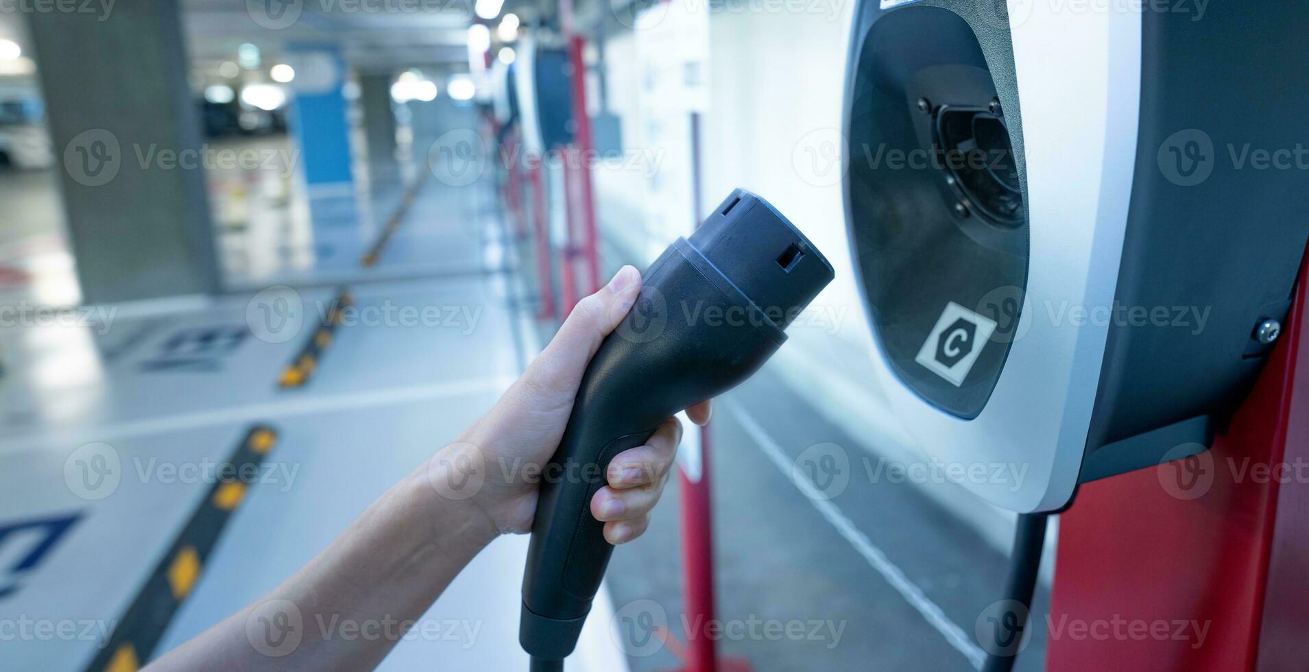 Hand holding electric charger at an electric vehicle charging station. Commercial EV car charging point for charge EV battery. Electric vehicle charging station at basement car parking lot of the mall photo