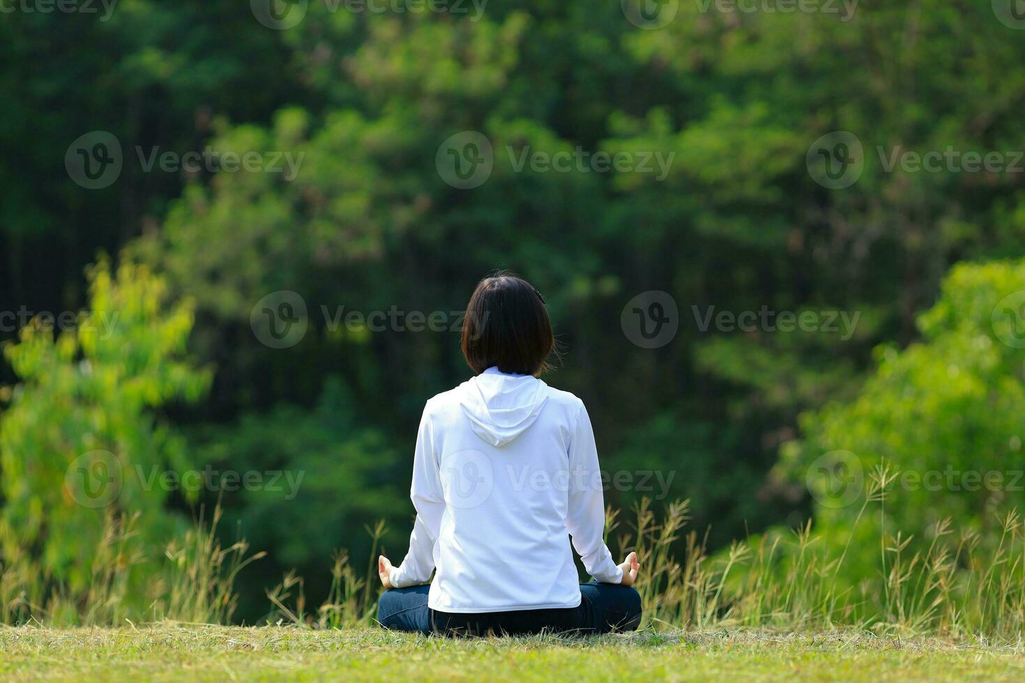 Rear view of woman in hoodie is relaxingly practicing meditation yoga in forest full of grass meadow in summer to attain happiness from inner peace wisdom with morning light for healthy mind and soul photo