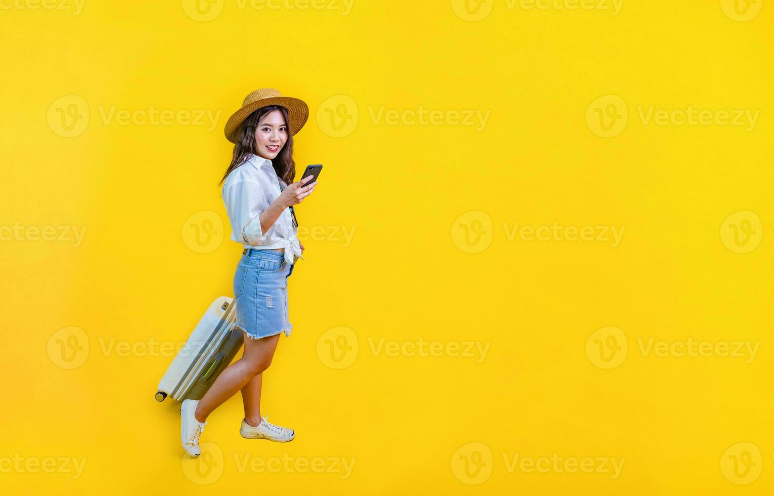 Pretty Asian woman passenger in trendy fashion is using mobile phone while carrying her luggage bag in happiness for travel and summer vacation isolated on yellow background photo