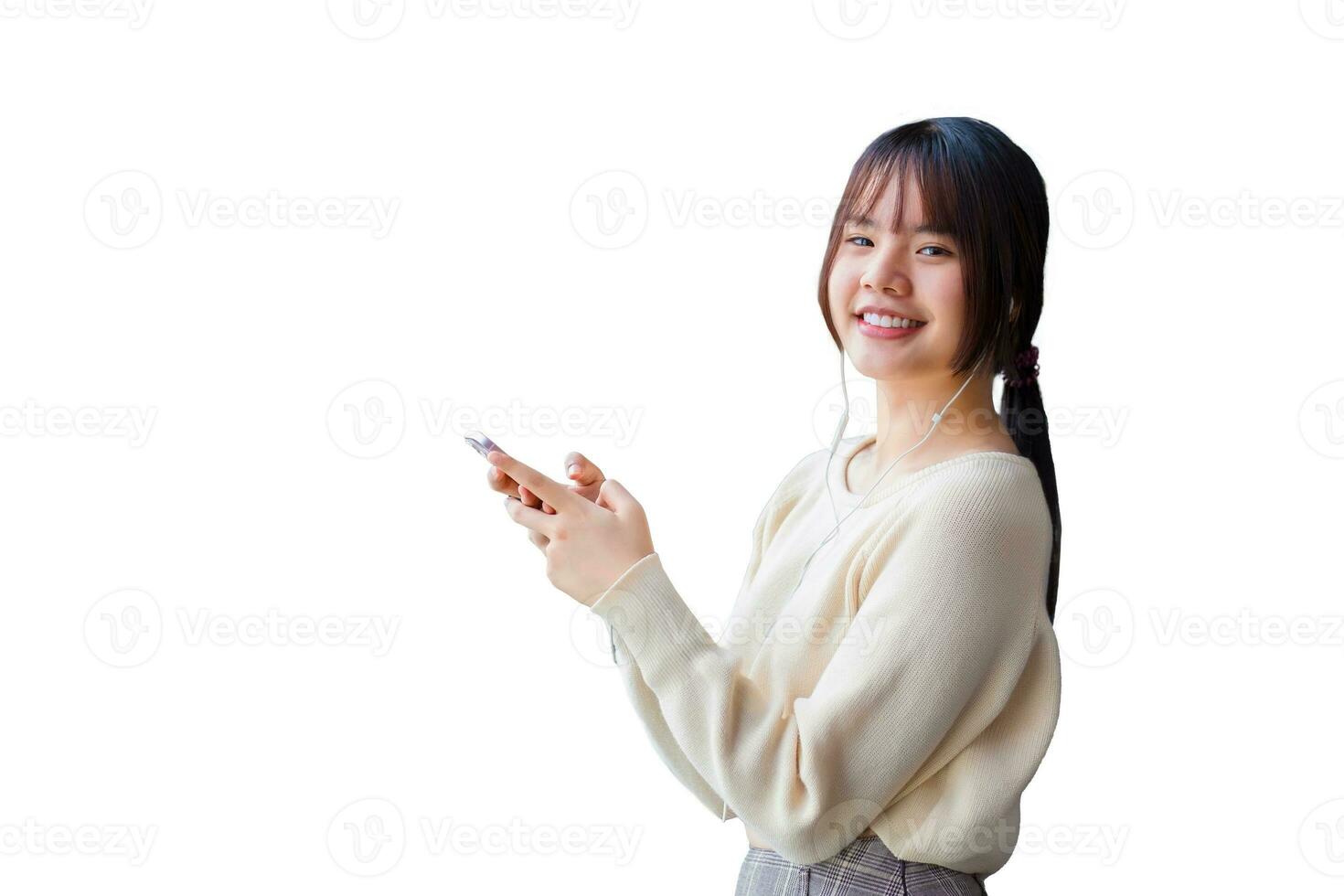 Cute young Asian student wearing long-sleeved shirt is standing and smiling while using her smartphone and headphones to listen lecture confidently while isolated on white background. photo