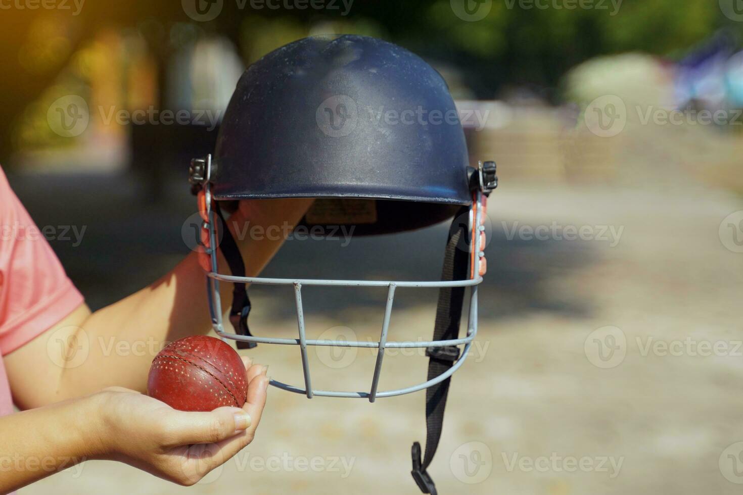 Cricket player holding cricket helmet and cricket ball in hands on the sidelines preparing before training on the field. Soft and selective focus. photo