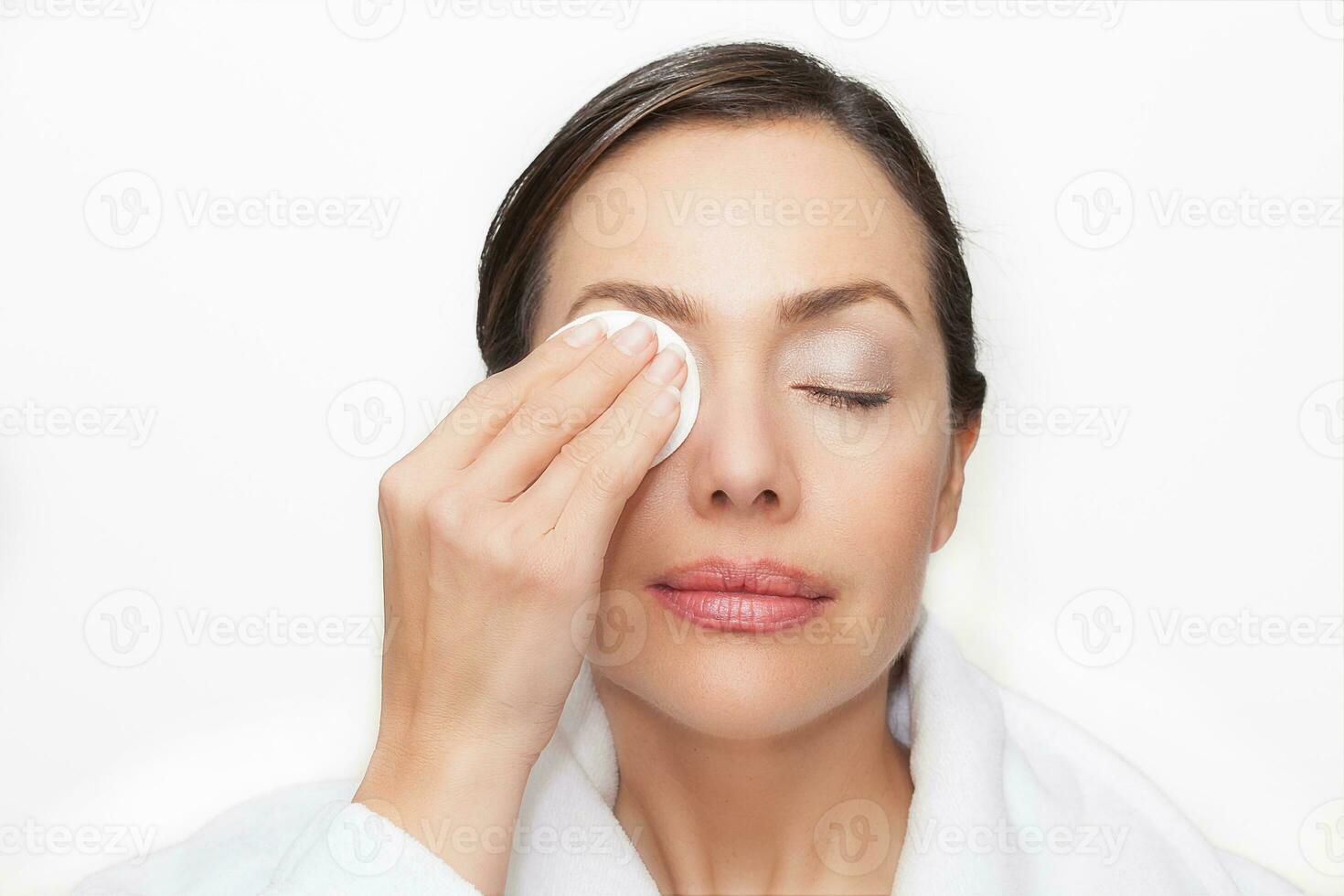 hermosa mujer quitando su maquillaje utilizando un algodón redondo vistiendo un blanco bata de baño terminado un blanco antecedentes foto