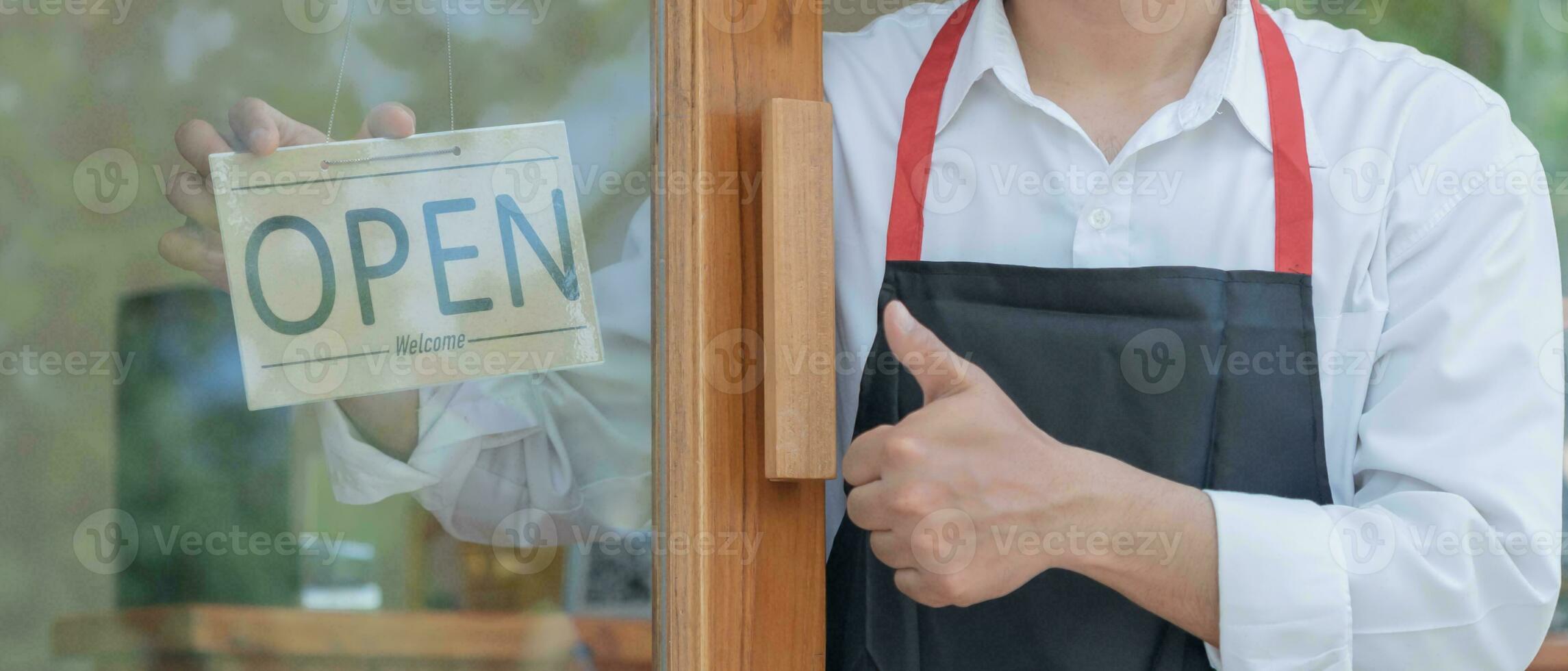 Business owner thumbs up to show his readiness to welcome customers. waitress man turning on open sign board on glass door in modern cafe coffee shop. New normal, small business, cafe, life style. photo
