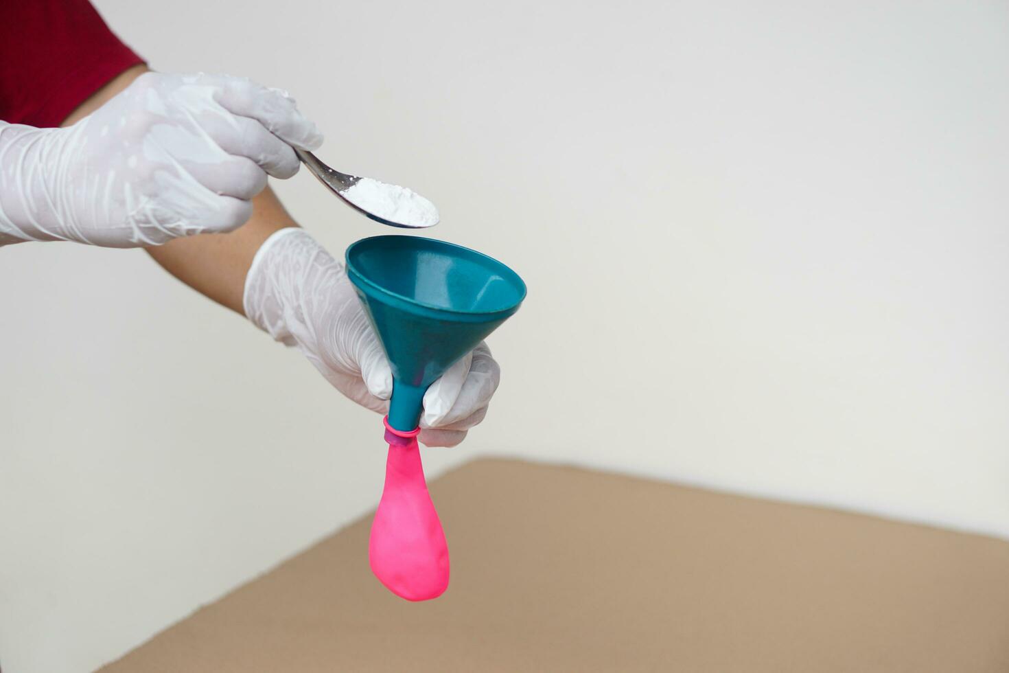 Closeup hands wears gloves holds funnel and flat pink balloon, put spoon of baking soda powder to pour into balloon. Concept, science experiment. photo