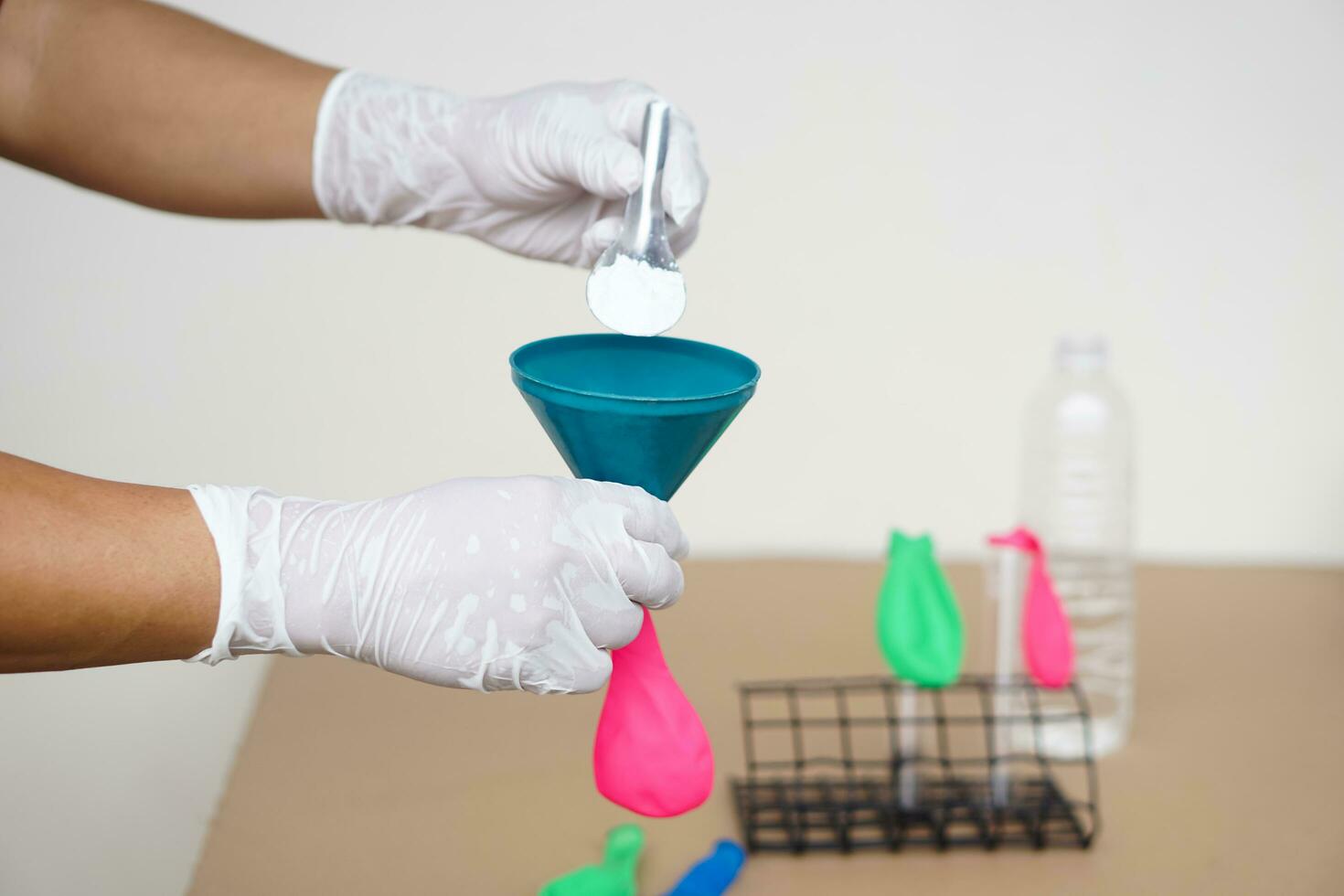 Closeup hands wears gloves holds funnel and flat pink balloon, put spoon of baking soda powder to pour into balloon. Concept, science experiment about reaction of chemical substance . photo