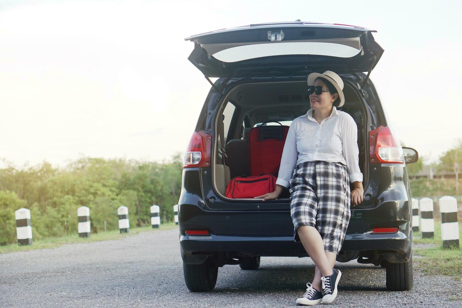 Happy middle aged Asian woman traveler sits at the trunk of car. Concept, relax time. Vacation or holiday trip by private vehicle. Transportation. Give time for yourself. photo