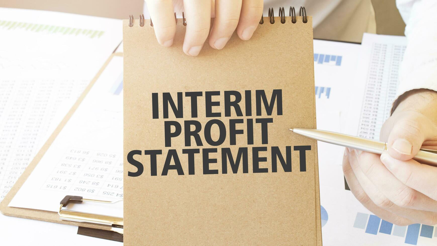 Text INTERIM PROFIT STATEMENT on brown paper notepad in businessman hands on the table with diagram. Business concept photo