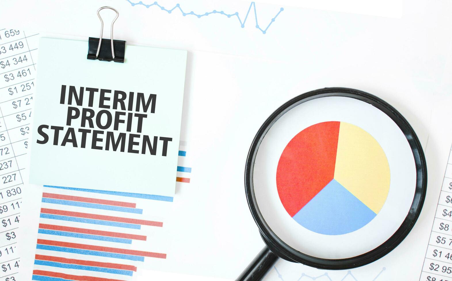 Green card on the white notepad and magnifier on the financial documentation. Text INTERIM PROFIT STATEMENT. Business concept photo