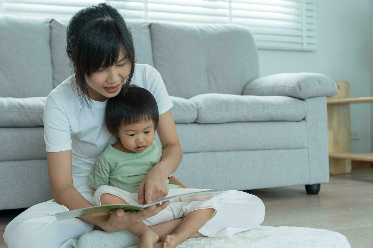 Happy Asian mother relax and read book with baby time together at home. parent sit on sofa with daughter and reading a story. learn development, childcare, laughing, education, storytelling, practice. photo