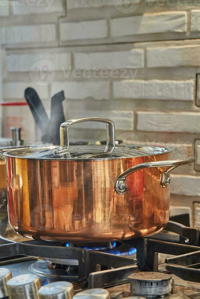 Copper saucepan with lid with dish is cooked over fire on gas stove. Steam rises above the pot photo