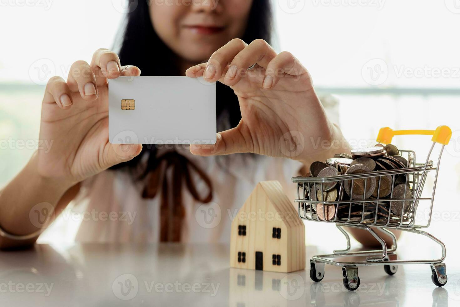 Selective focus of hand holding a blank credit chip card with wooden house model and full of coins in shopping cart photo