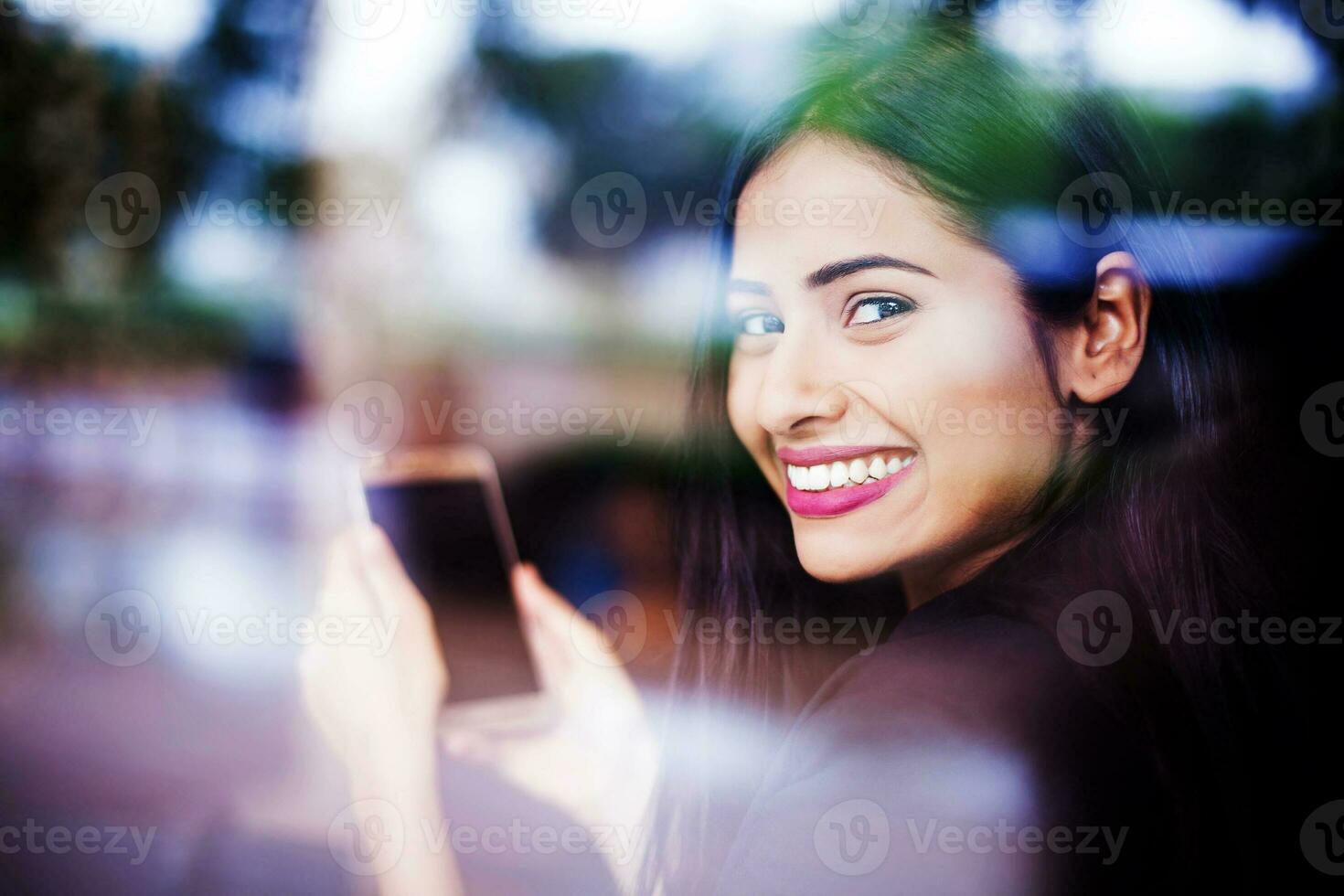 A beautiful young Indian woman smiling and looking through a glass window while holding a phone with blank screen in her hands photo