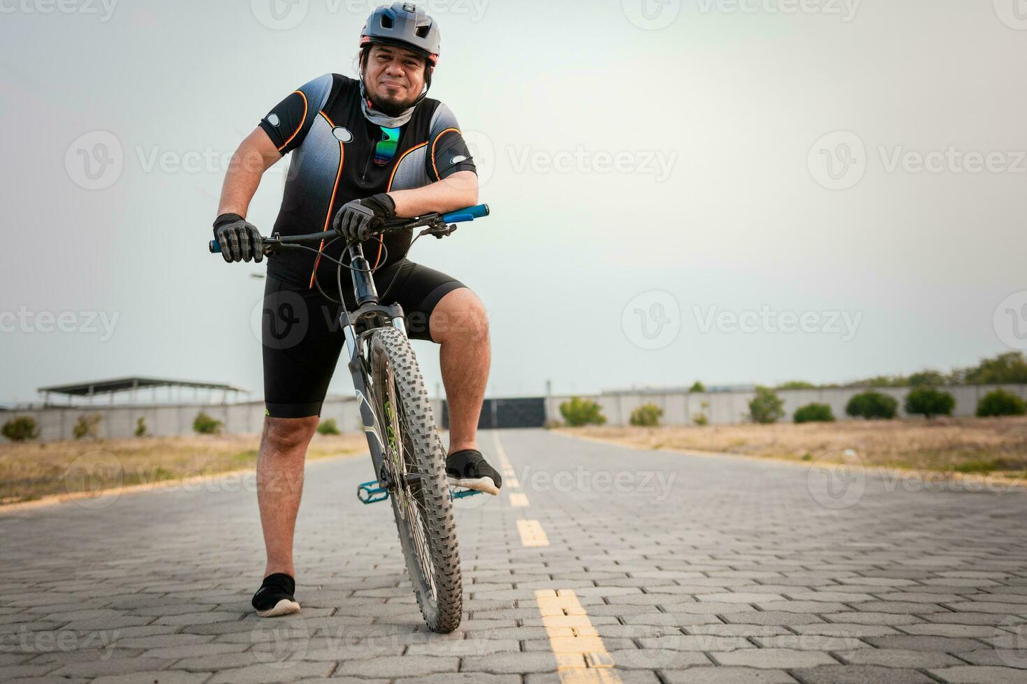 sonriente ciclista en su bicicleta mirando a el cámara en el la carretera.  gordito masculino ciclista en ropa de deporte montando un bicicleta al aire  libre 26416263 Foto de stock en Vecteezy