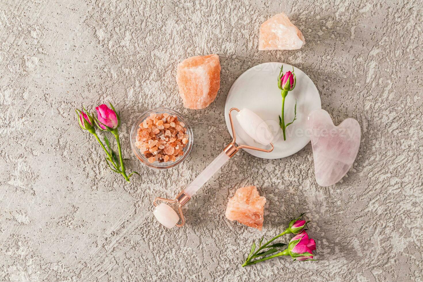 Top view of quartz roller and gua sha on a white round marble stand and rosebuds and lumps of salt. The concept of spa, relaxation. cement background. photo