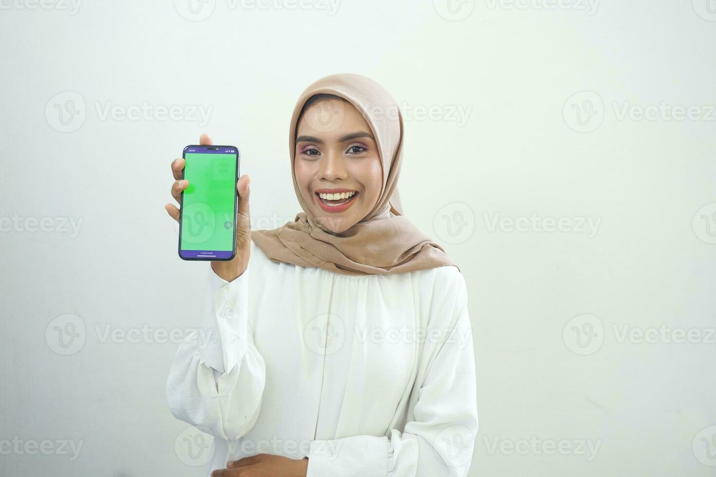 Excited beautiful Asian muslim woman showing green screen mobile phone isolated over white background photo