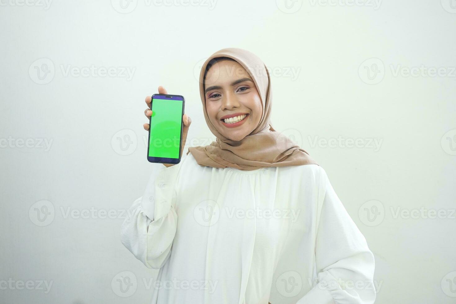 Excited beautiful Asian muslim woman showing green screen mobile phone isolated over white background photo