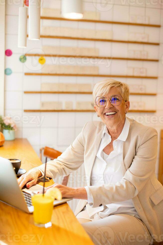 Senior woman using mobile phone while working on laptop and drinking fresh orange juice in the cafe photo
