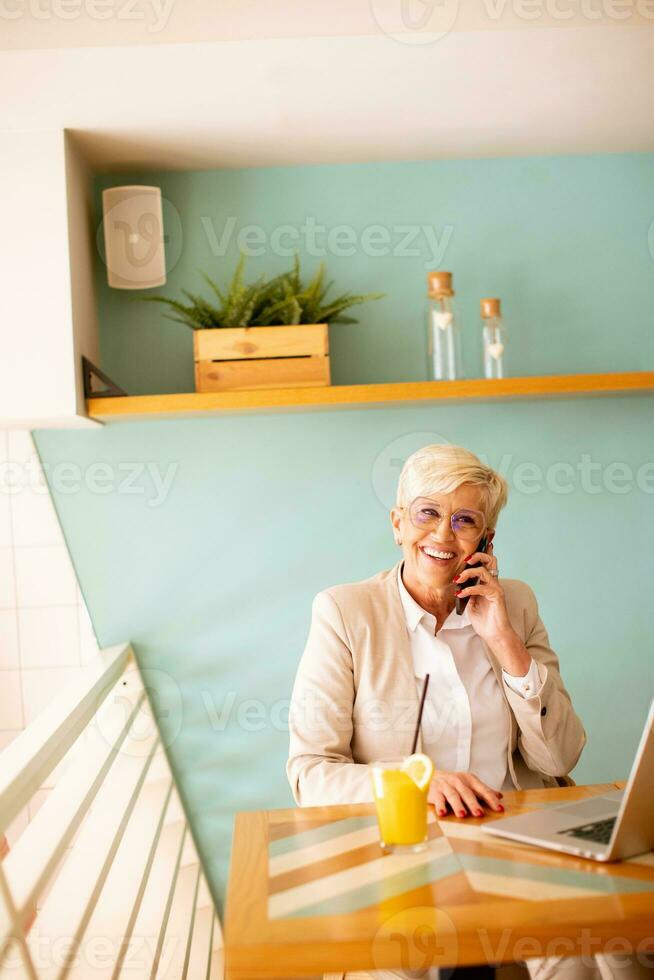 Senior woman using mobile phone while working on laptop and drinking fresh orange juice in the cafe photo