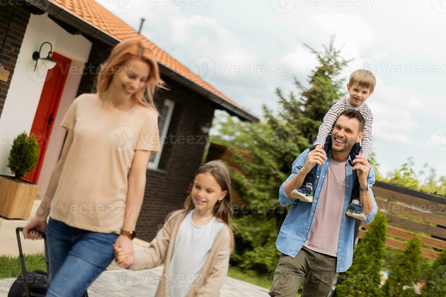 Family with a mother, father, son and daughter walking with baggage outside on the front porch of a brick house photo