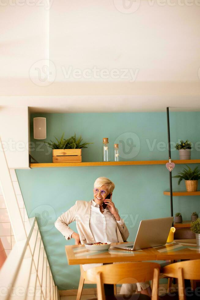 Senior woman using mobile phone while working on laptop and having healthy breakfast in the cafe photo