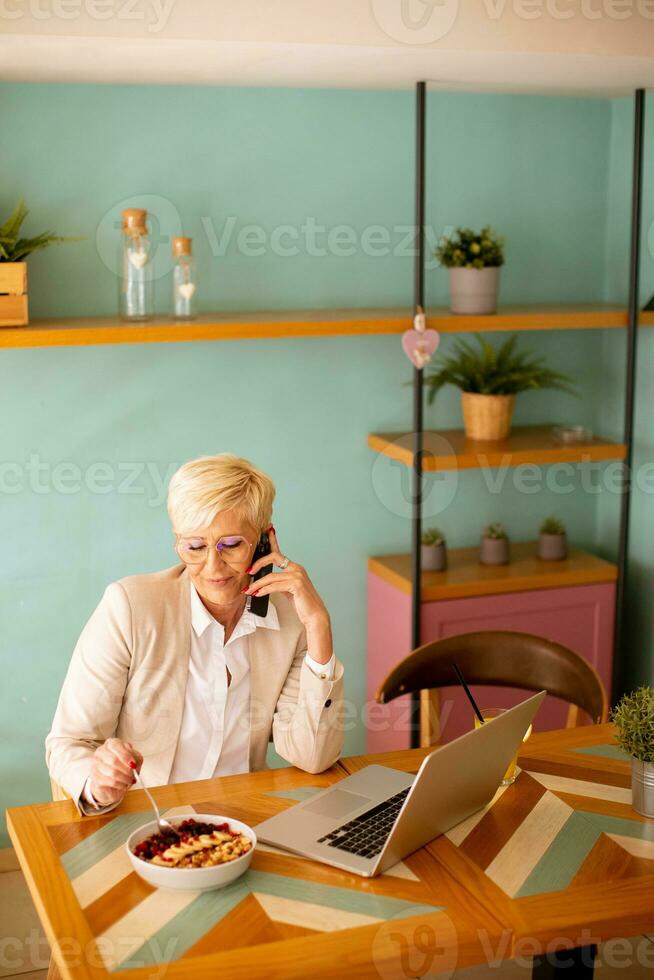 Senior woman using mobile phone while working on laptop and having healthy breakfast in the cafe photo