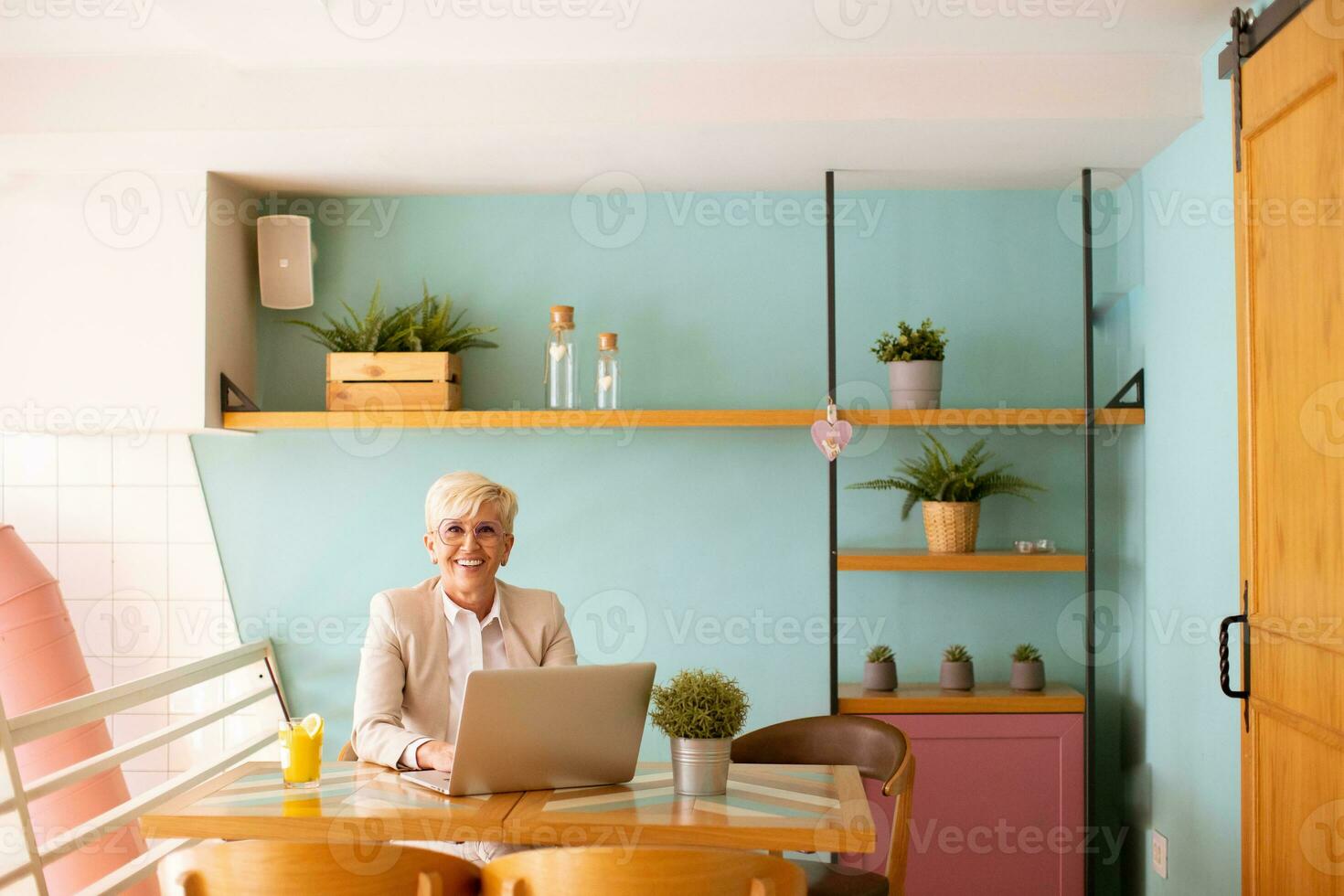 Senior woman using mobile phone while working on laptop and drinking fresh orange juice in the cafe photo