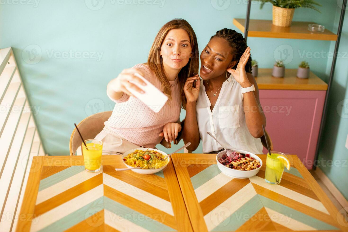 Two young women, caucasian and black one, taking selfie with mobile phone in the cafe photo