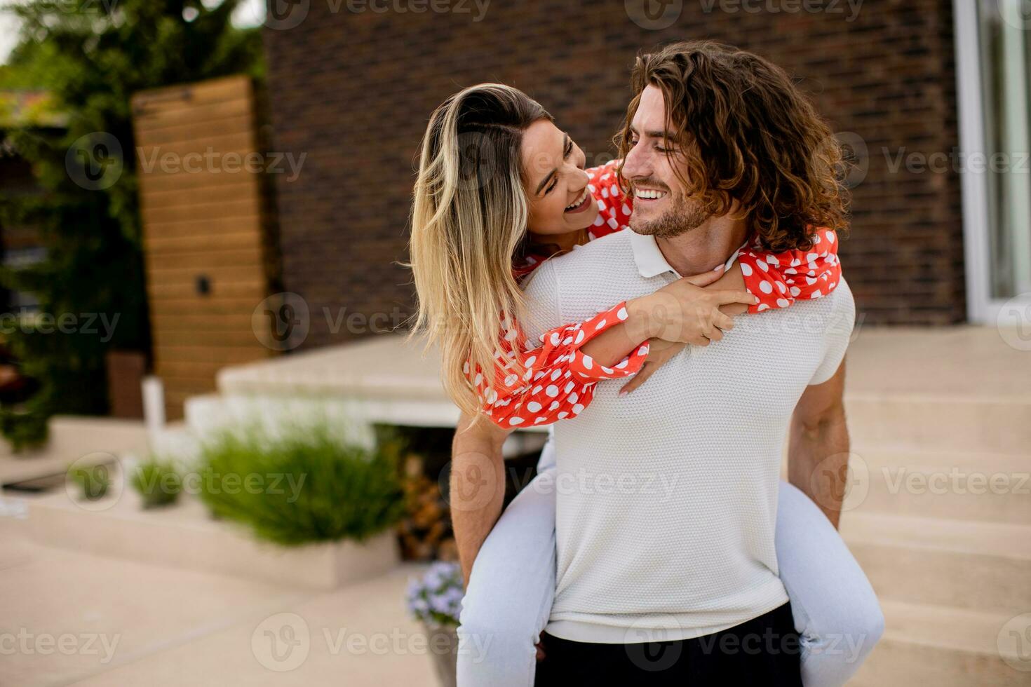 Handsome long hair man carrying the young woman on his back in front of brick house photo