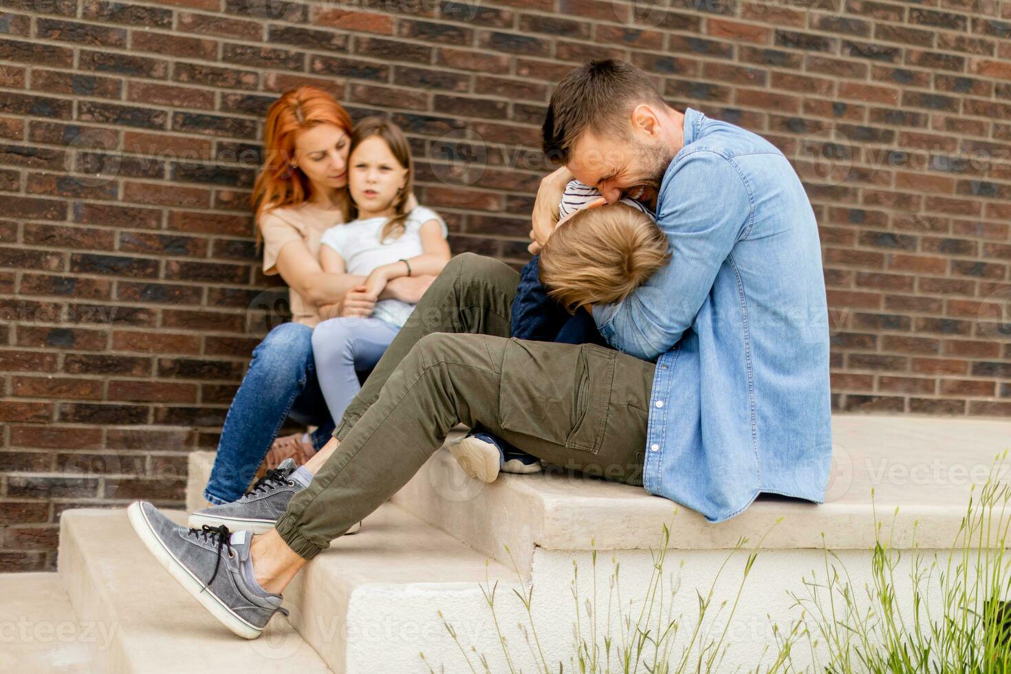 familia con un madre, padre, hijo y hija sentado fuera de en el pasos de un frente porche de un ladrillo casa foto