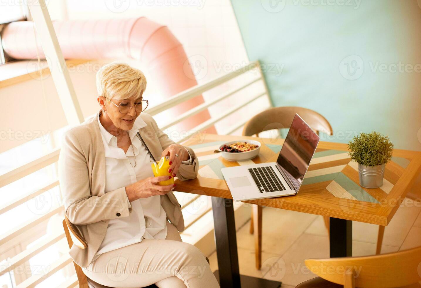 Senior woman using mobile phone while working on laptop and drinking fresh orange juice in the cafe photo