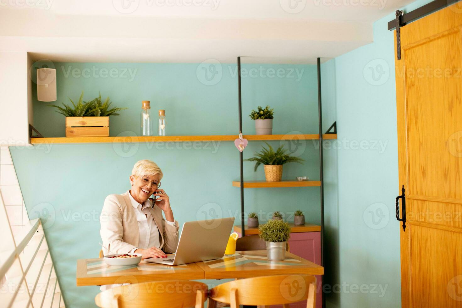Senior woman using mobile phone while working on laptop and drinking fresh orange juice in the cafe photo