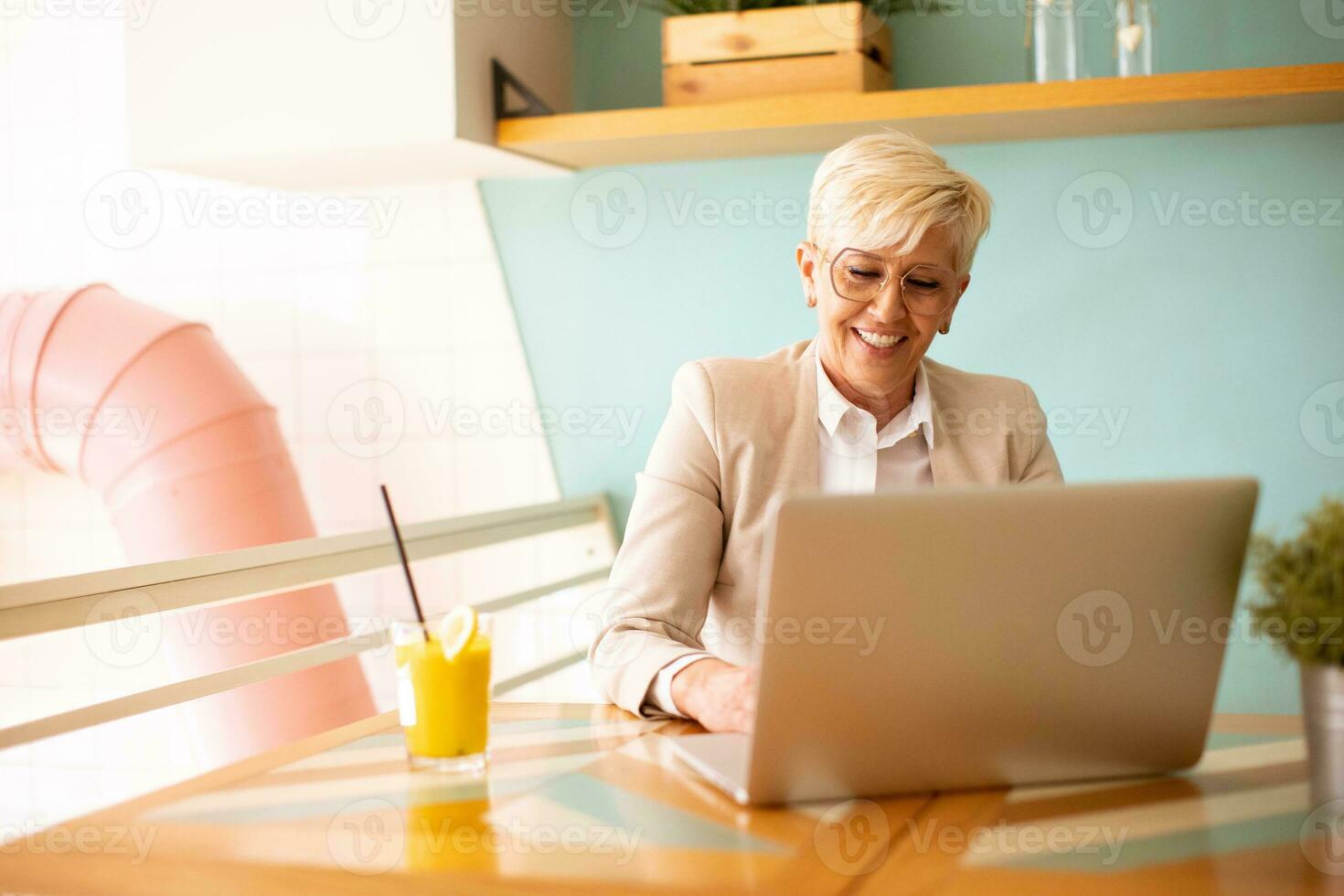 Senior woman using mobile phone while working on laptop and drinking fresh orange juice in the cafe photo