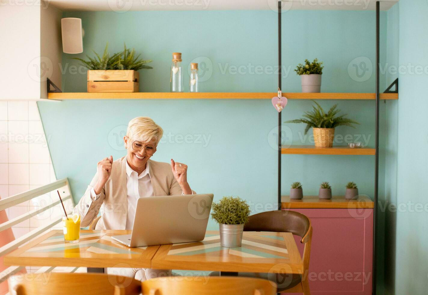 Senior woman using mobile phone while working on laptop and drinking fresh orange juice in the cafe photo