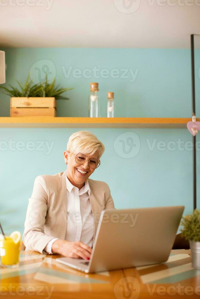 Senior woman using mobile phone while working on laptop and drinking fresh orange juice in the cafe photo