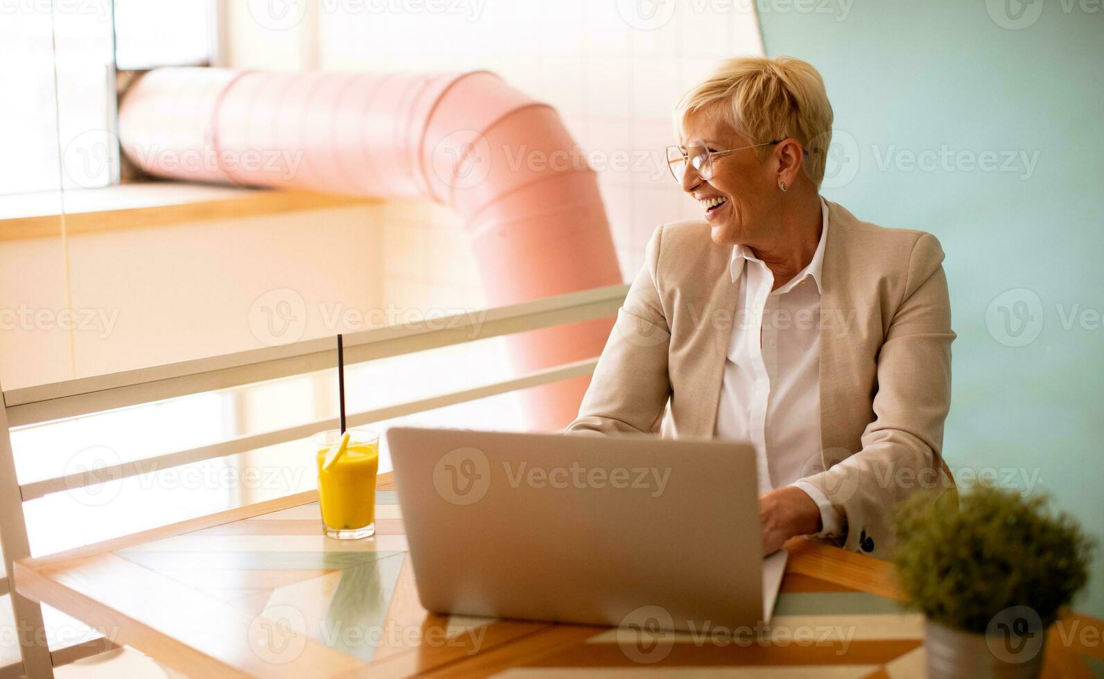 Senior woman using mobile phone while working on laptop and drinking fresh orange juice in the cafe photo