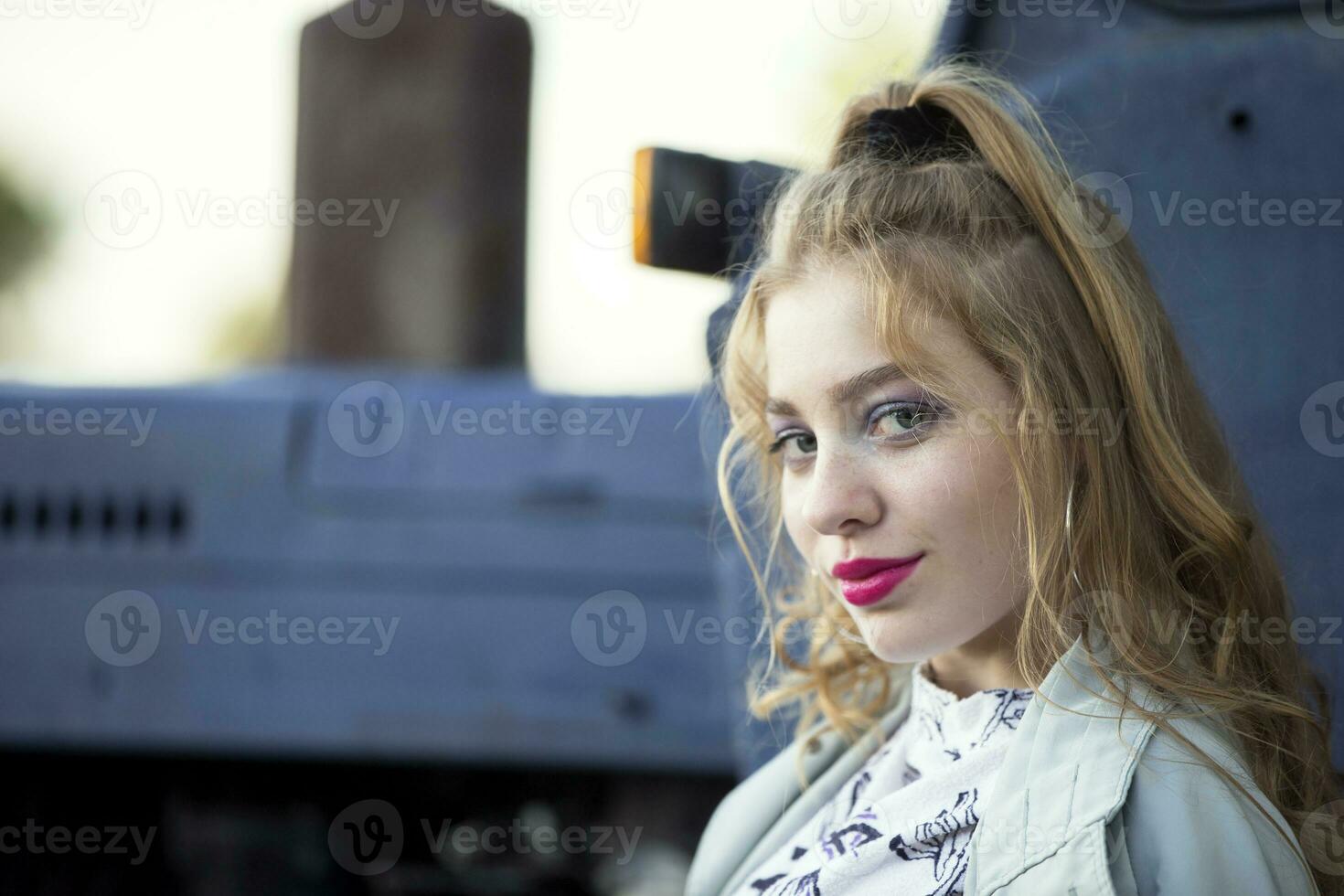 A beautiful country girl in the style of the 90s in bright clothes stands near a tractor. photo