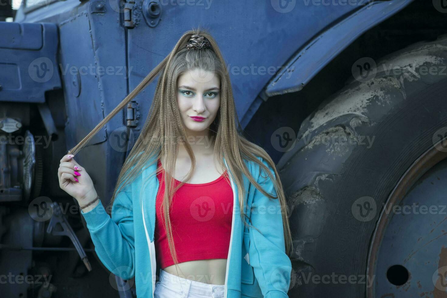 A beautiful country girl in the style of the 90s in bright clothes stands near a tractor. photo