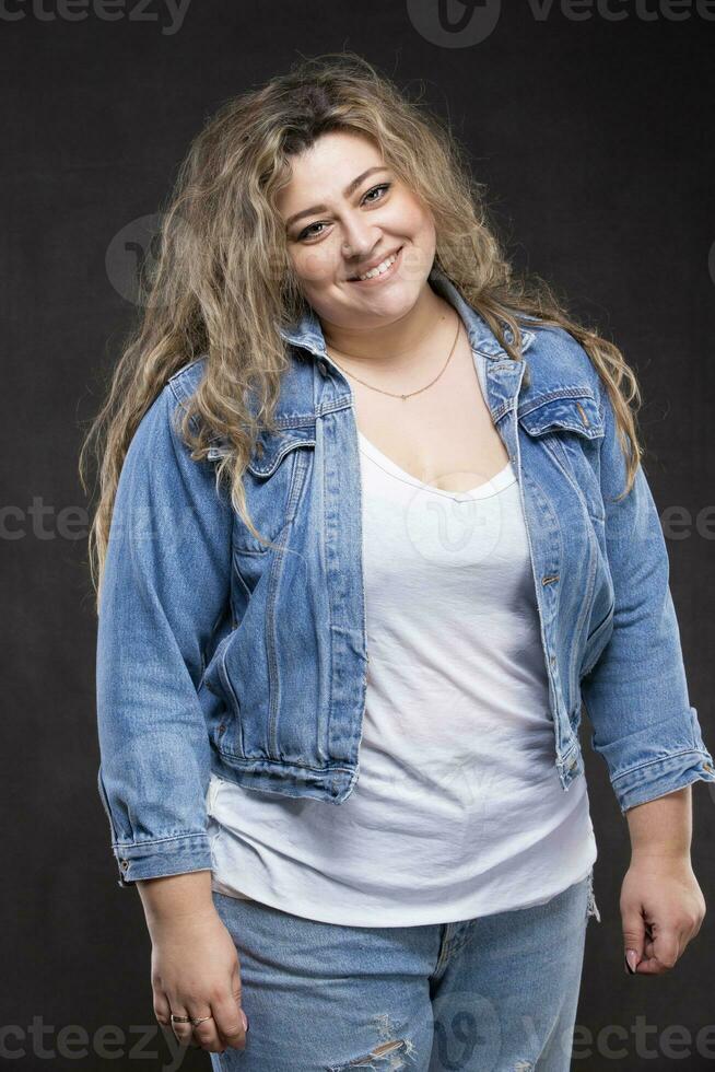 un hermosa grasa mujer en un mezclilla chaqueta mira a el cámara y sonrisas en un gris antecedentes. foto