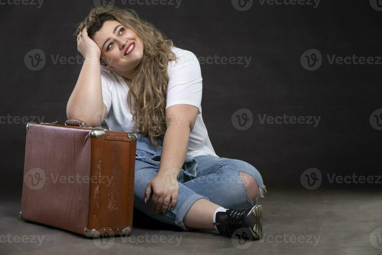 Beautiful fat woman in denim clothes with an old retro suitcase on a gray background. Plus size model girl. photo