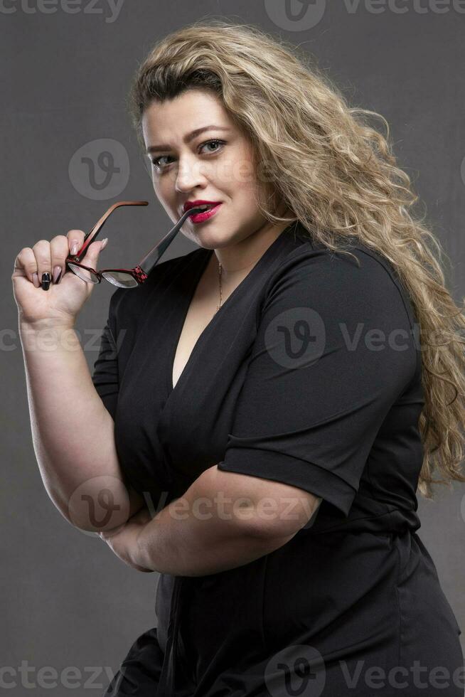 un hermosa grasa mujer con largo Rizado pelo sostiene lentes en su manos seguras de sí mismas rechoncho mujer. foto